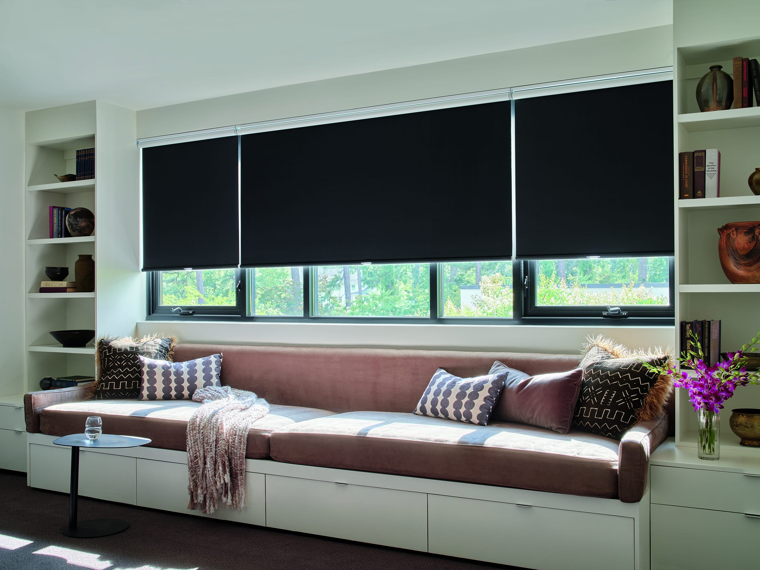 Three black blackout roller shades lowered over a long window above a built in window seat