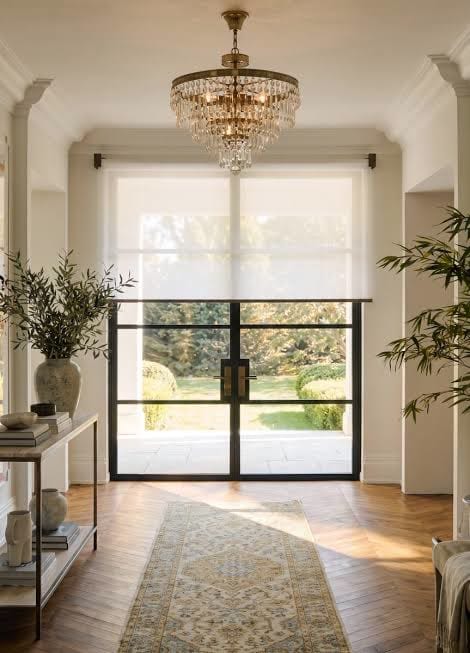 A sheer white roller shade drawn half way over black framed glass doors in a bright entrance hall